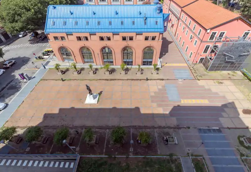 Aerial view of a paved plaza with a central statue, adjacent brick buildings and planted tree beds; bright daylight and clear architectural details.