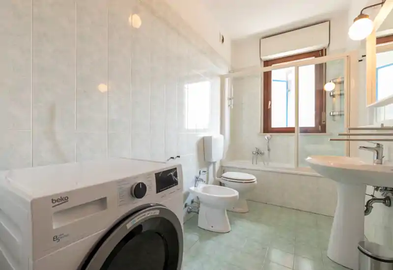 Bright, clean bathroom with bathtub-shower, pedestal sink, toilet, bidet and a washing machine near the entrance; natural light from a window keeps the space airy.
