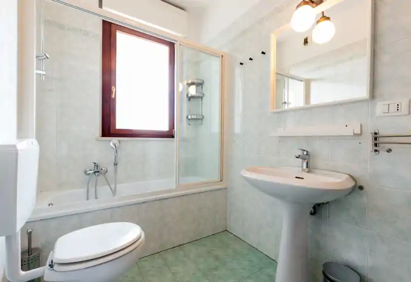 Clean, compact bathroom with a bathtub and glass shower screen, pedestal sink, toilet and a large window providing natural light.