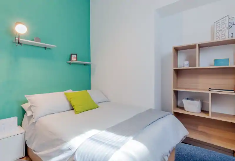 Bright, neatly styled bedroom with a queen bed against a teal accent wall, soft linens and a wooden shelving unit. Natural light and minimal decor create a calm, inviting space ideal for listings.