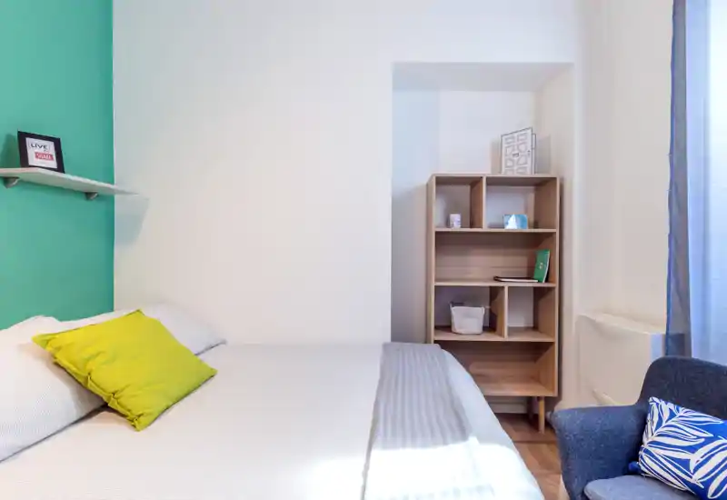 Bright, modern bedroom with a neatly made bed, colorful cushions and a wooden shelving unit; natural light from a window creates a fresh, welcoming atmosphere.