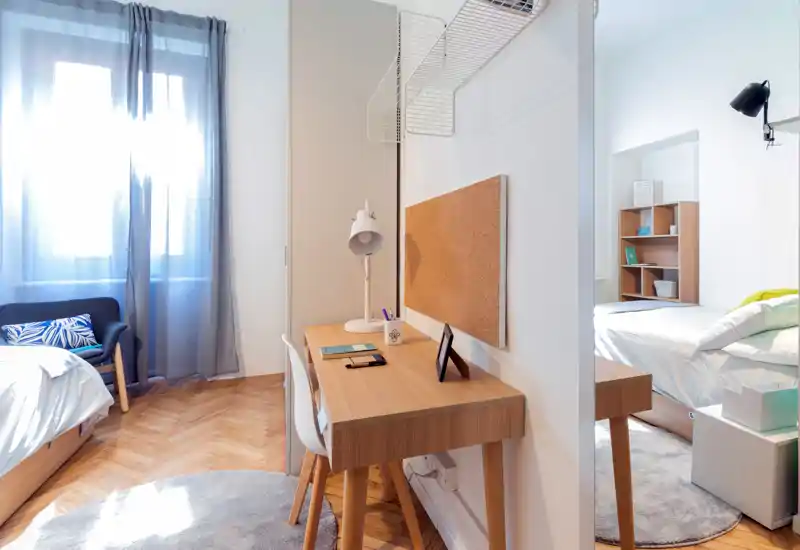 Bright, well-styled bedroom with a wooden desk, cozy rugs and natural light streaming from a large window — ideal for studying or relaxing.