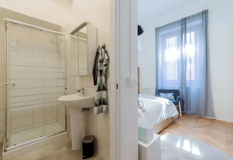 Bright, simply styled bedroom visible through a doorway with a comfortable bed, armchair and large window letting in natural light.