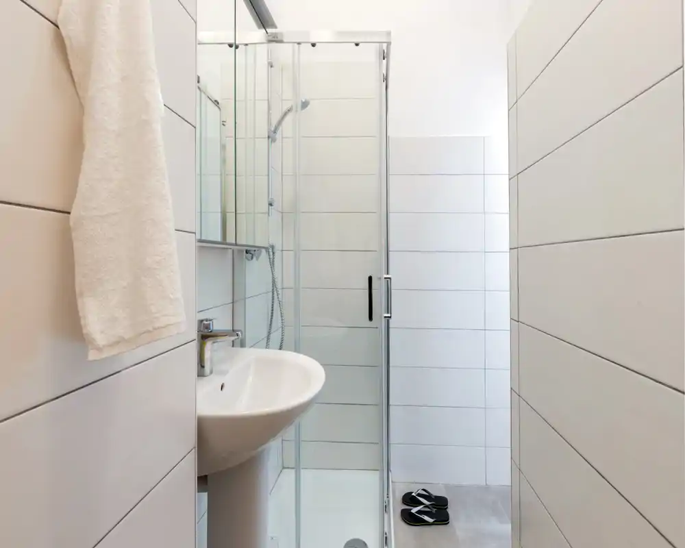 Salle de bains compacte et moderne avec lavabo sur colonne, douche à paroi vitrée et carrelage neutre — lumineuse et propre, parfaite pour les locations courte durée.