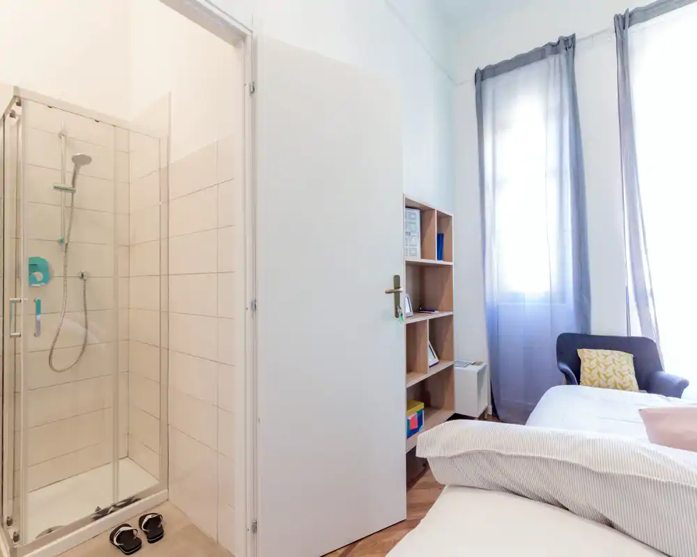 Chambre lumineuse et compacte avec lit visible, fauteuil et étagères ; lumière naturelle à travers des voilages et une petite salle d'eau avec douche visible par la porte ouverte.
