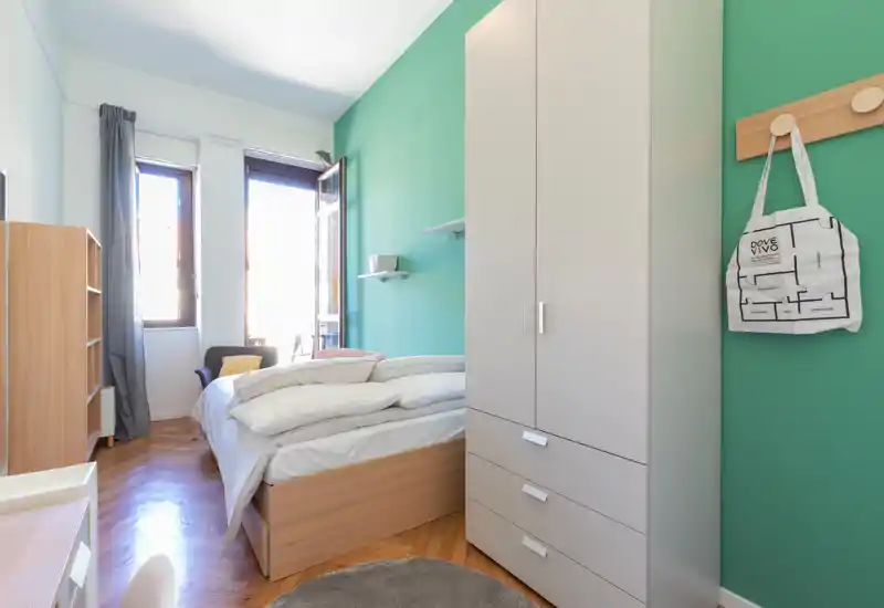 Chambre lumineuse et soignée avec un lit double, parquet et une grande armoire contre un mur vert-bleu. De larges fenêtres inondent la pièce de lumière naturelle et les textiles apportent une atmosphère chaleureuse.