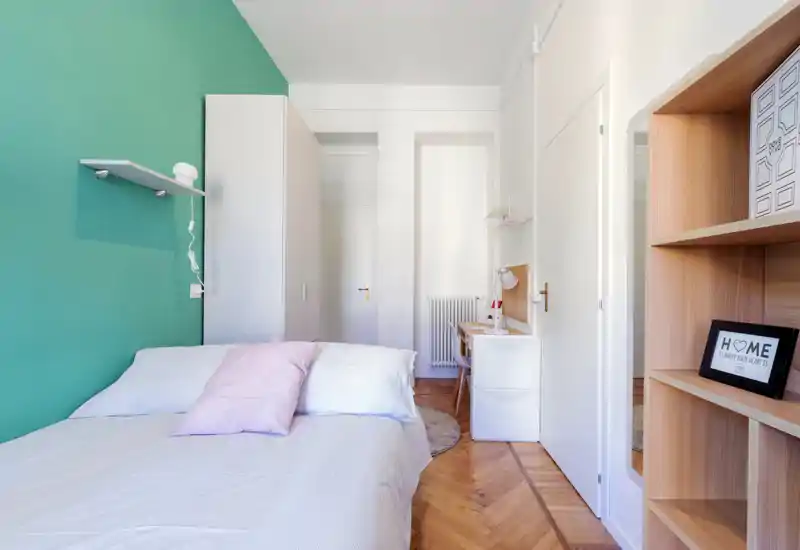 Chambre moderne et lumineuse avec un lit double bien fait, des accents pastel et un mur d'accent turquoise. La pièce est bien éclairée, aménagée avec des étagères et un bureau pour une ambiance cosy et compacte.