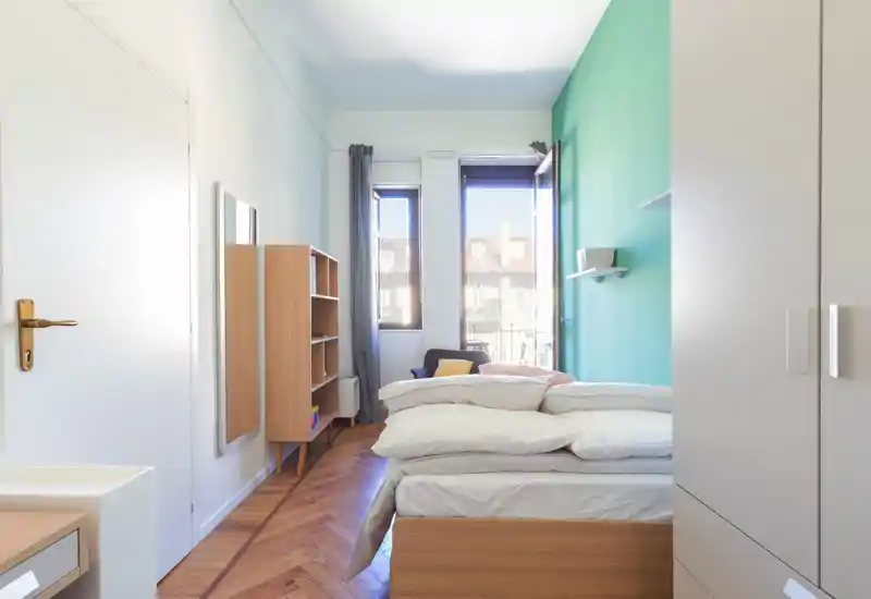 Chambre moderne et lumineuse avec lit à cadre bois, literie blanche superposée et lumière naturelle provenant de deux hautes fenêtres ; coussins décoratifs et un fauteuil près de la fenêtre.