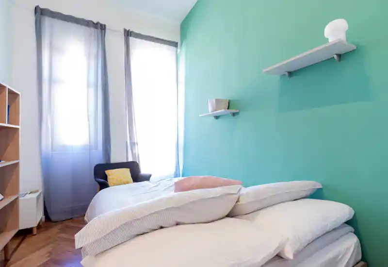 Chambre lumineuse et moderne avec un lit double bien fait contre un mur bleu-vert, de grandes fenêtres aux voilages légers et un fauteuil confortable — idéale pour mettre en avant la lumière naturelle et une décoration contemporaine.