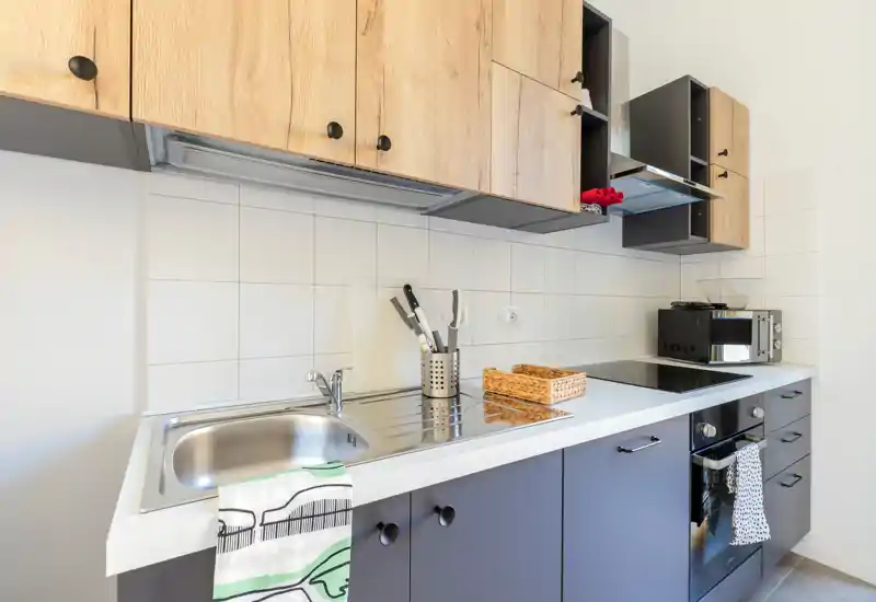 Cuisine compacte et moderne avec placards supérieurs effet bois, éléments bas foncés, évier inox, plaque à induction et four encastré — soigneusement agencée avec des ustensiles et un petit panier en osier.