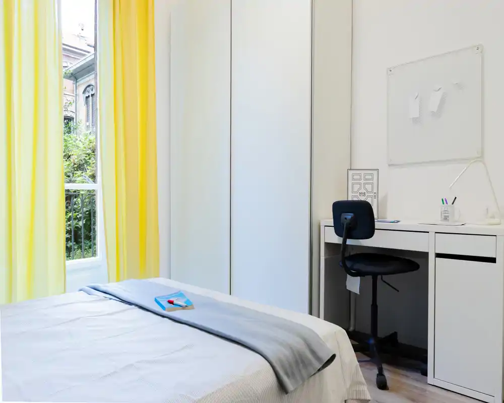 Dormitorio moderno y luminoso con cama bien hecha, armario blanco y pequeño escritorio junto a la ventana. Las cortinas amarillas y la luz natural crean un ambiente fresco y acogedor, perfecto para alquileres temporales.