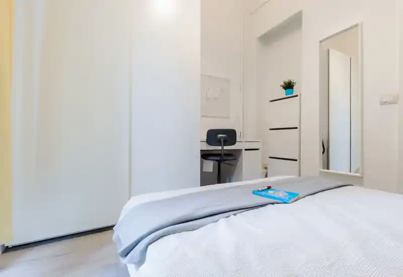 Dormitorio moderno y luminoso con la cama bien hecha en primer plano, un escritorio compacto con silla y un espejo de cuerpo entero. Tonos neutros y decoración simple que transmiten una sensación limpia y acogedora.