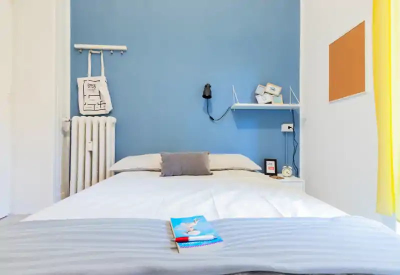 Dormitorio luminoso y ordenado con pared azul, ropa de cama blanca y un cojín decorativo; bien iluminado por luz natural y decoración moderna y minimalista.