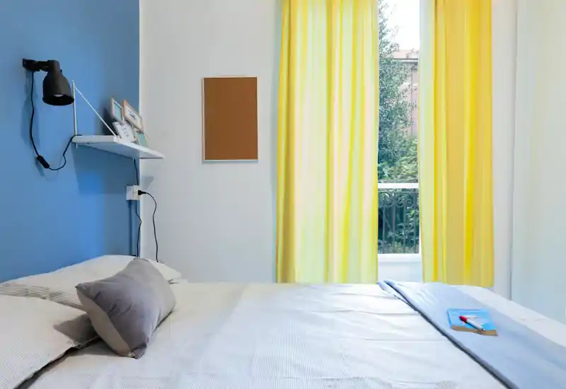 Dormitorio luminoso y bien arreglado con cama amplia, ropa de cama suave y ventana con cortinas amarillas que aportan luz natural.