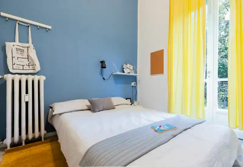 Dormitorio luminoso y con estilo con una cama doble frente a una pared azul, ropa de cama suave, una lámpara en la estantería y una gran ventana con cortinas amarillas que deja entrar mucha luz natural.