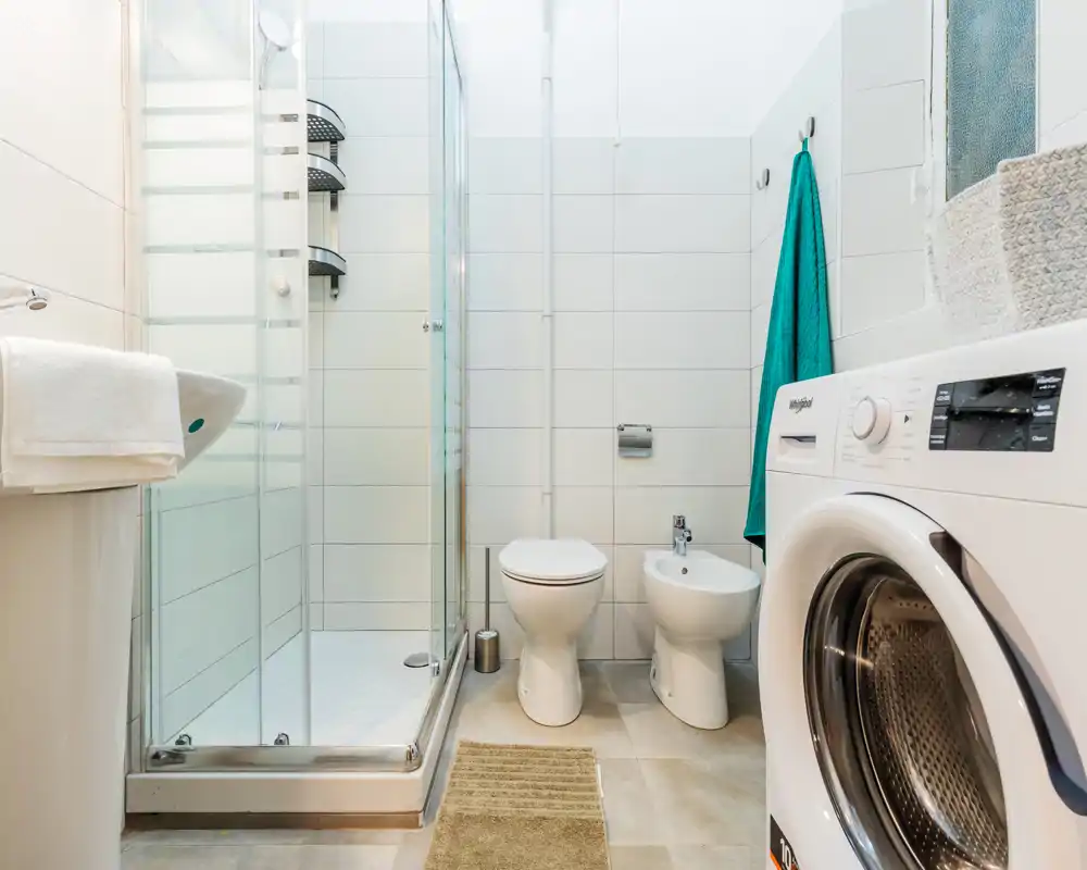 Baño moderno y luminoso con cabina de ducha, inodoro, bidé y lavadora. Azulejos blancos y suelo neutro crean un espacio limpio y práctico, perfecto para alquileres temporales.
