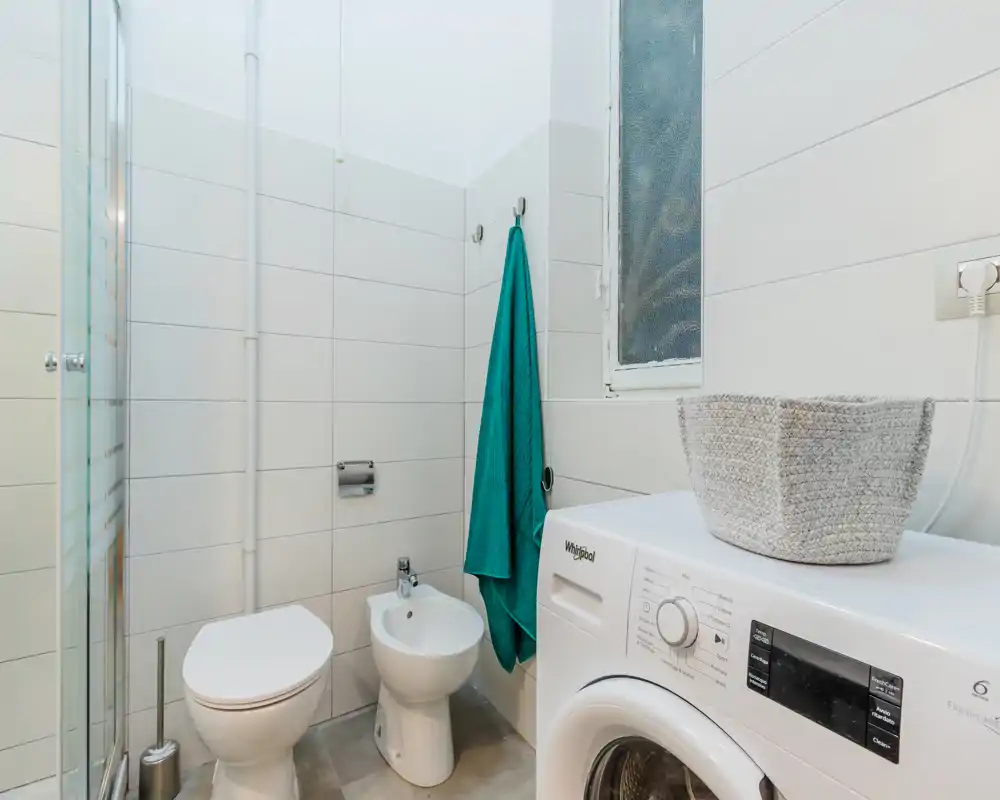 Baño compacto y limpio con inodoro, bidé y lavadora; azulejos blancos y ventana esmerilada que aportan buena luz.