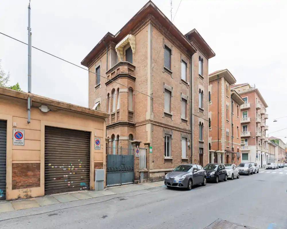 Vista de la calle con un edificio residencial de ladrillo, coches aparcados y puertas de garaje; fotografía exterior diurna que destaca la fachada y el entorno urbano.