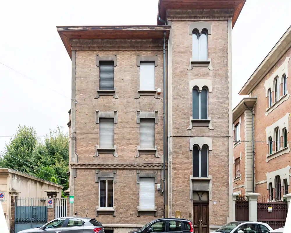 Vista exterior de un edificio residencial de ladrillo con persianas cerradas, ventanas en arco y coches aparcados delante — adecuada como foto de fachada para un anuncio.