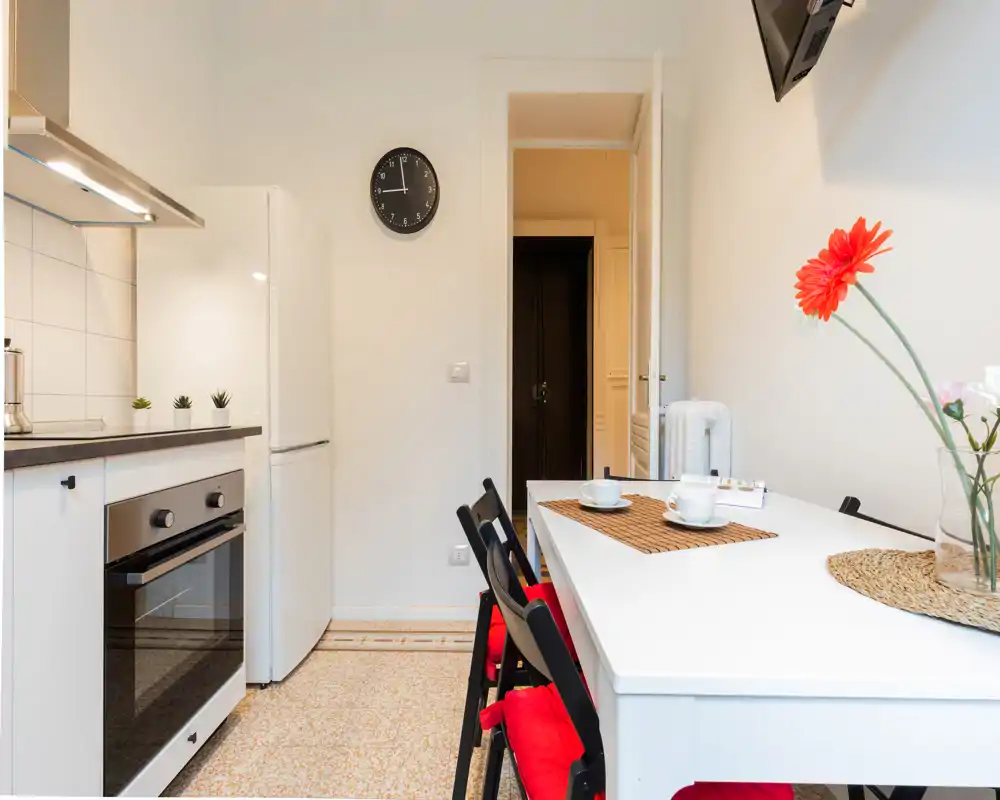 Cocina compacta y luminosa con mesa blanca y sillas con cojines rojos, horno y nevera modernos y detalles decorativos sencillos (flores y manteles), ideal para un piso pequeño.