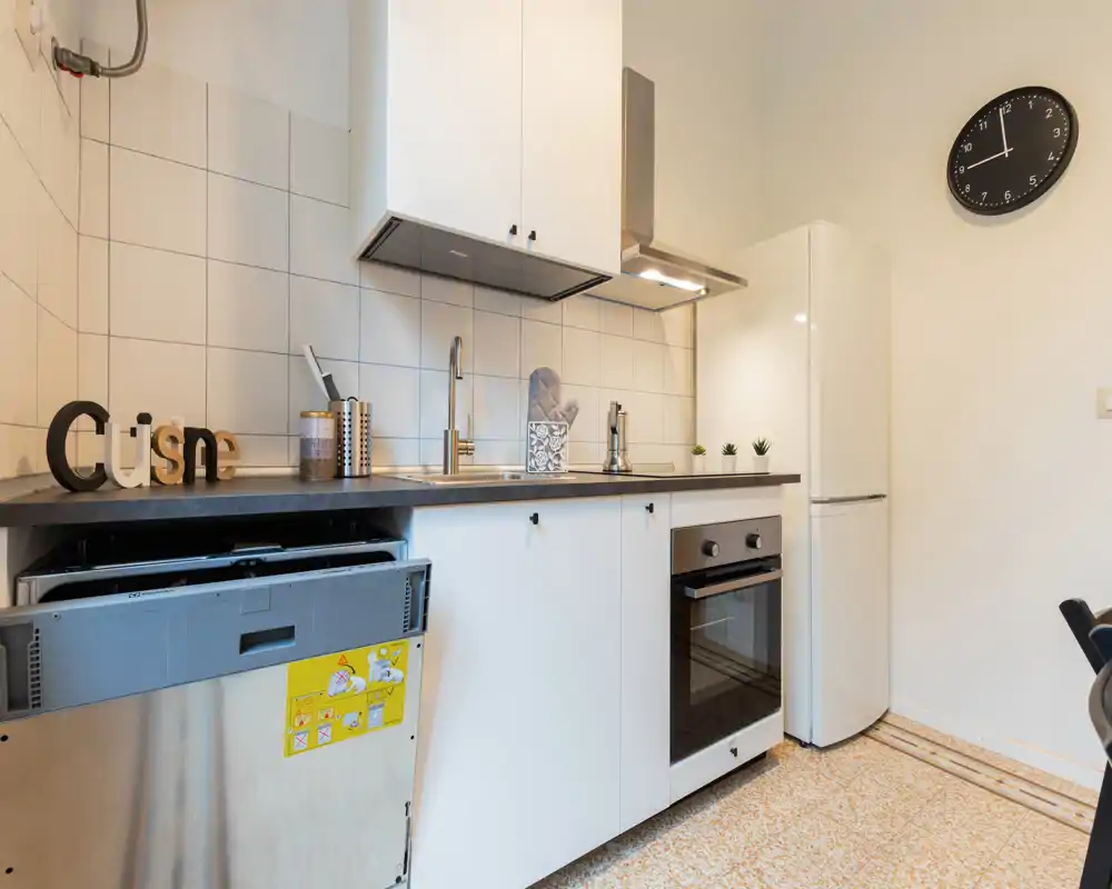 Cocina moderna y compacta con muebles blancos, horno empotrado, frigorífico y fregadero de acero; encimera ordenada con detalles decorativos y buena iluminación.