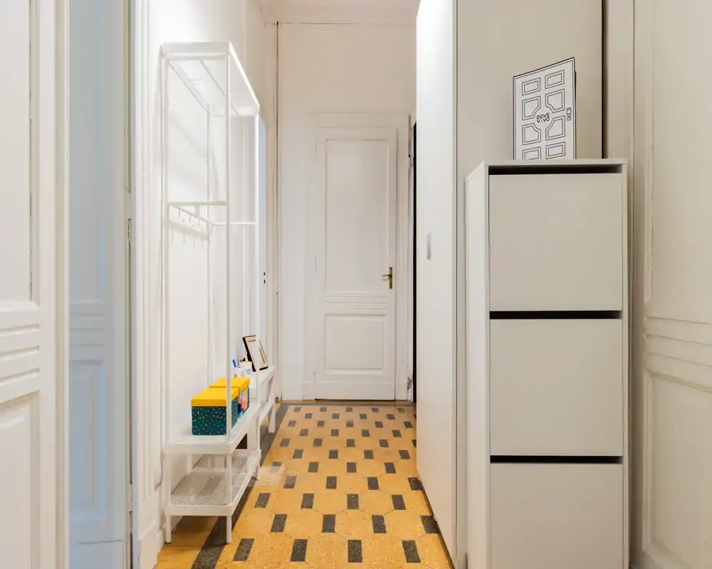 Entrada luminosa y ordenada con zapatero estrecho y perchero abierto, suelo de baldosa decorada y puerta blanca al fondo — ideal para causar una buena primera impresión del piso.