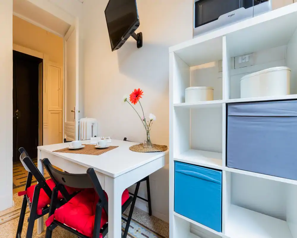 Pequeña zona de comedor con mesa blanca, dos sillas plegables negras con cojines rojos, individuales y un jarrón con flores. Iluminado, ordenado y bien preparado para un apartamento pequeño.