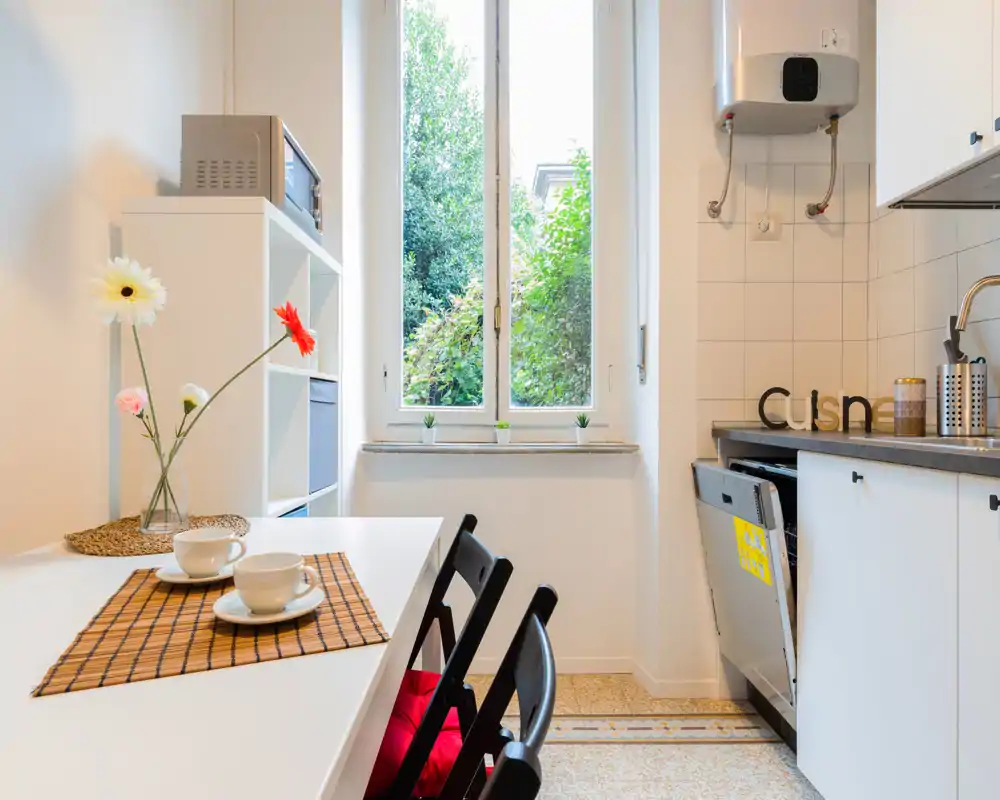 Cocina luminosa y compacta con mesa junto a la ventana, muebles modernos y flores decorativas que crean un ambiente acogedor.