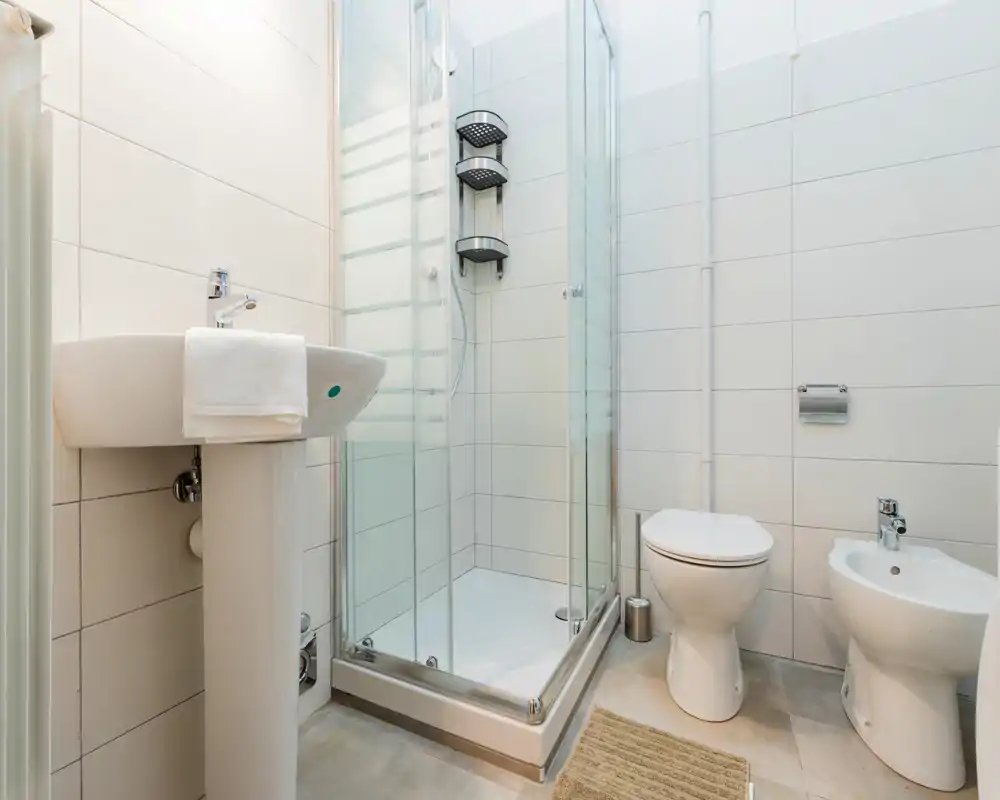 Baño moderno y luminoso con ducha de cristal, lavabo de pie, inodoro y bidé; azulejos neutros y accesorios limpios que ofrecen un espacio funcional.