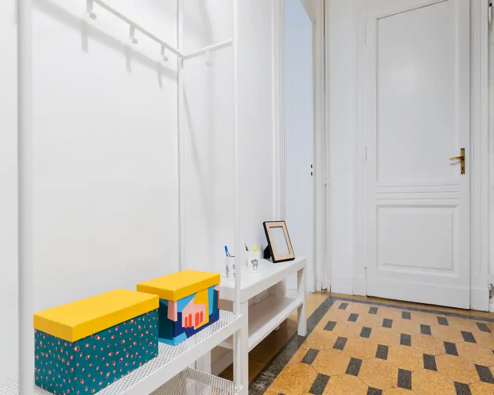 Entrada luminosa y ordenada con perchero blanco, banco y cajas decorativas sobre un suelo de baldosas con patrón.
