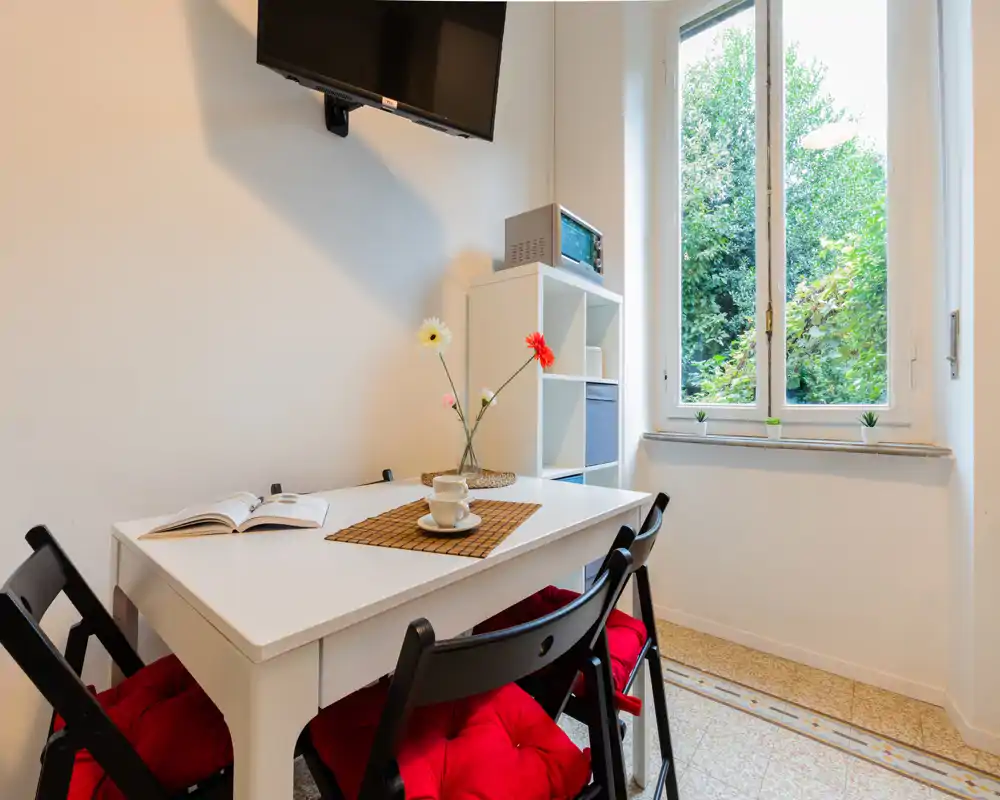 Acogedor rincón comedor bien iluminado con mesa blanca, cuatro sillas negras con cojines rojos, flores decorativas y una ventana con luz natural.