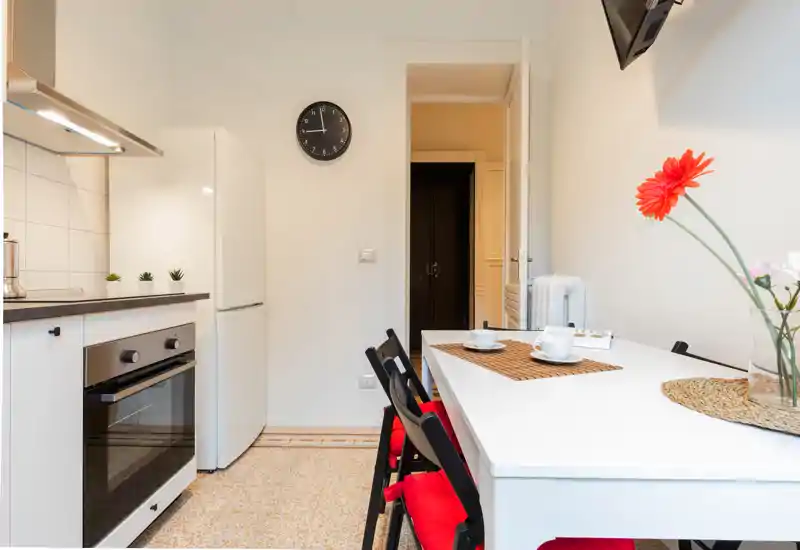 Bright, compact kitchen with a white table and red-cushioned chairs, modern oven and fridge, and simple decorative touches (flowers and placemats) ideal for a small apartment.