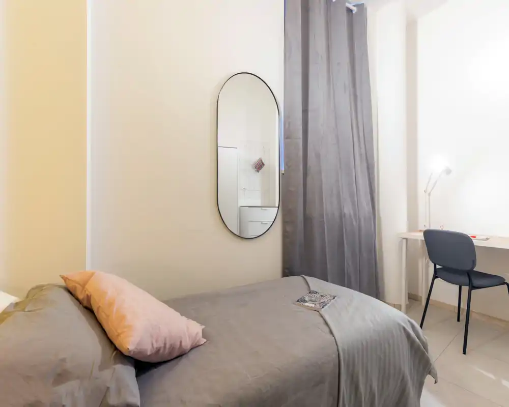 Dormitorio luminoso y ordenado con cama en tonos neutros, cojín decorativo, espejo de pared y un pequeño escritorio junto a la ventana — perfecto para descansar y trabajar.