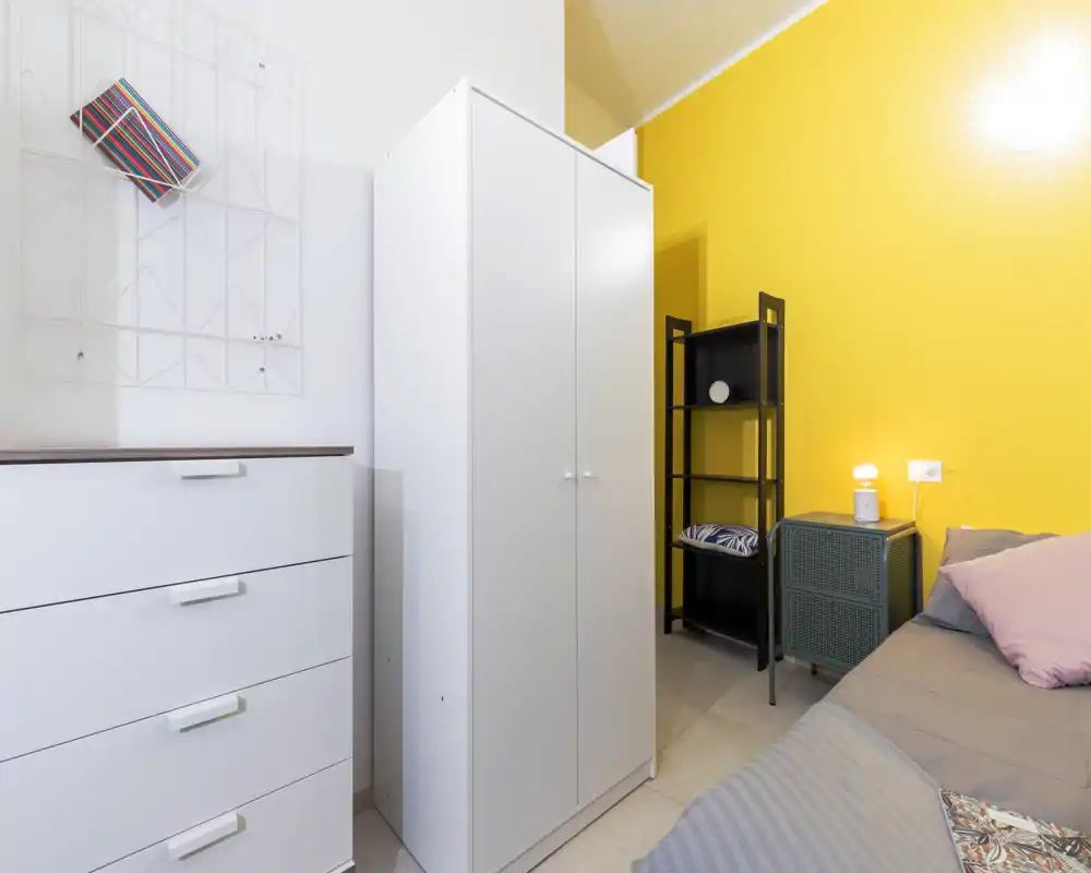 Rincón de dormitorio compacto y luminoso con cama gris y cojines pastel, armario y cómoda blancos y una pared amarilla intensa que aporta personalidad.