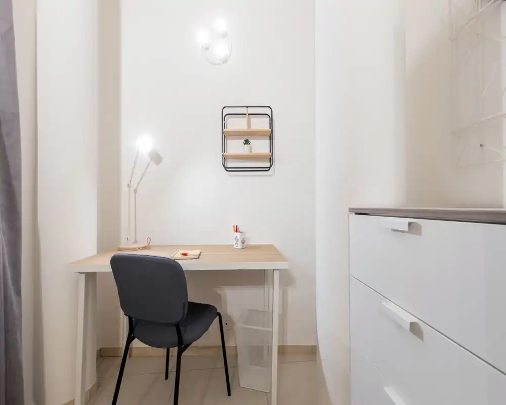 Rincón de trabajo luminoso y minimalista con escritorio sencillo, lámpara y silla. Paredes blancas y una pequeña estantería en la pared crean un espacio de trabajo funcional y ordenado.