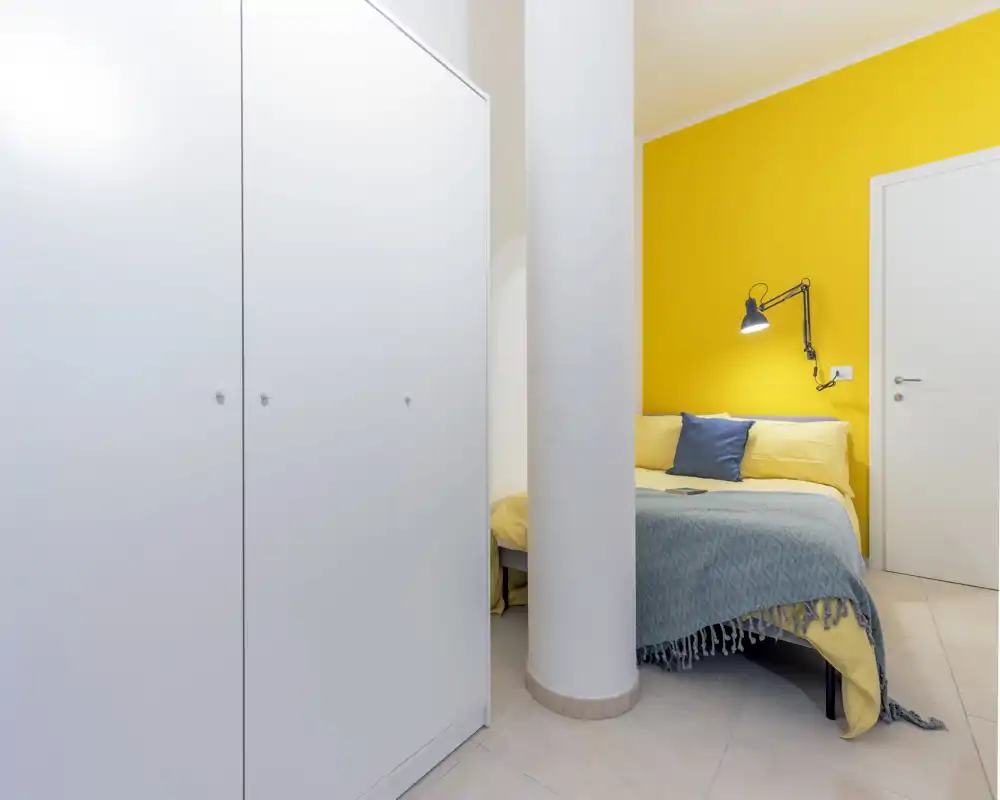 Bright, cozy bedroom with a yellow accent wall, modular wardrobe and a neatly made bed with cushions and a throw — ideal for rental photos highlighting cheerful decor.