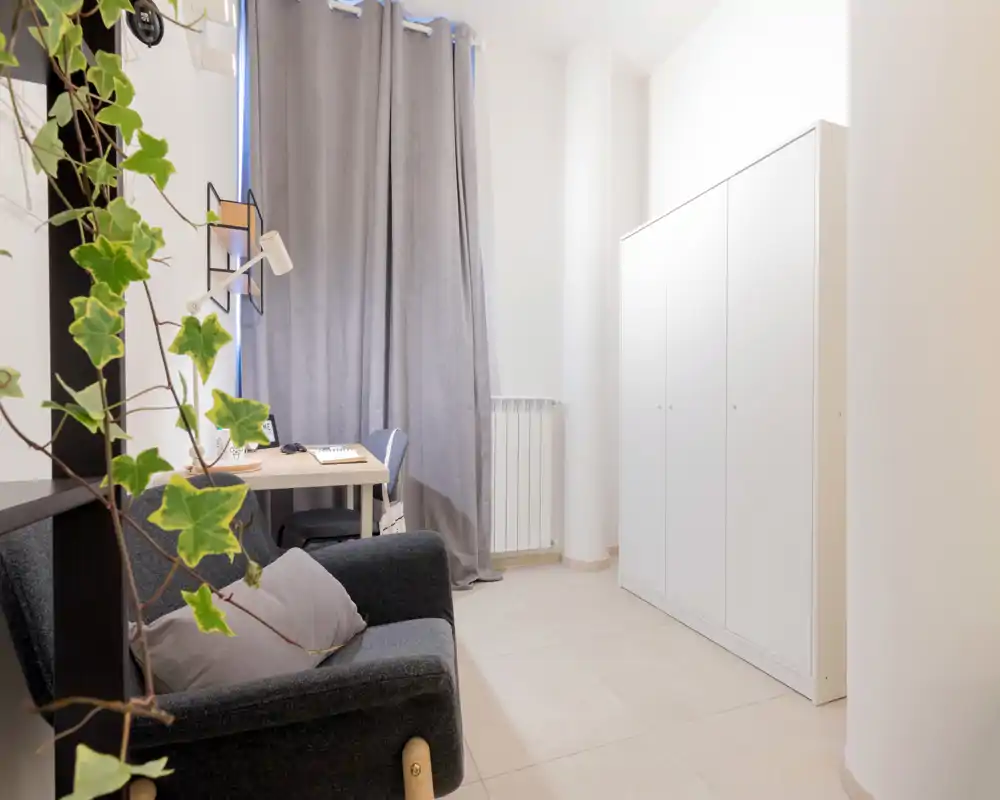 Cozy small living area with a dark upholstered sofa, a desk by the curtained window and a white wardrobe; neutral tones and plants add a calm, modern feel.
