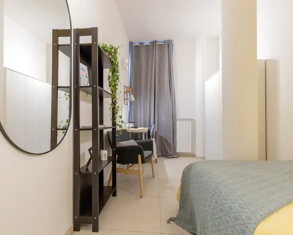 Bright, modern bedroom with a visible bed covered by a patterned throw, a shelving unit, armchair and a small desk by the window — clean minimal styling and soft neutral tones.