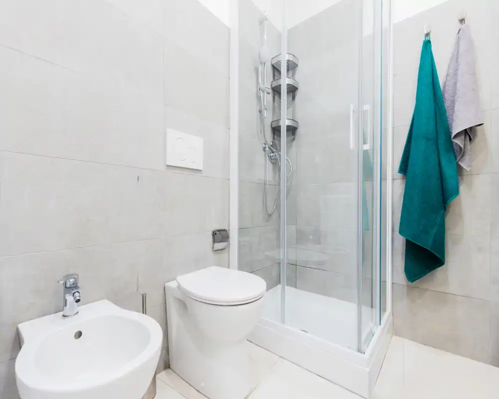Modern, compact bathroom with a shower enclosure, toilet and bidet set against light stone tiles; fresh towels hang on the wall for a clean, guest-ready feel.