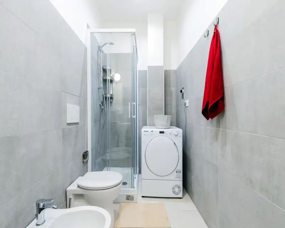Modern, compact bathroom with tiled walls, glass shower cabin, toilet, bidet and a washer/dryer unit. Bright, clean styling ideal for apartment listings.