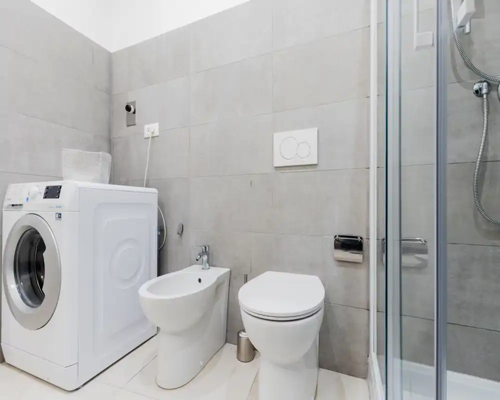 Modern bathroom with a shower, toilet, bidet and a washing machine; light grey tiles and bright, clean finishes give a fresh look.