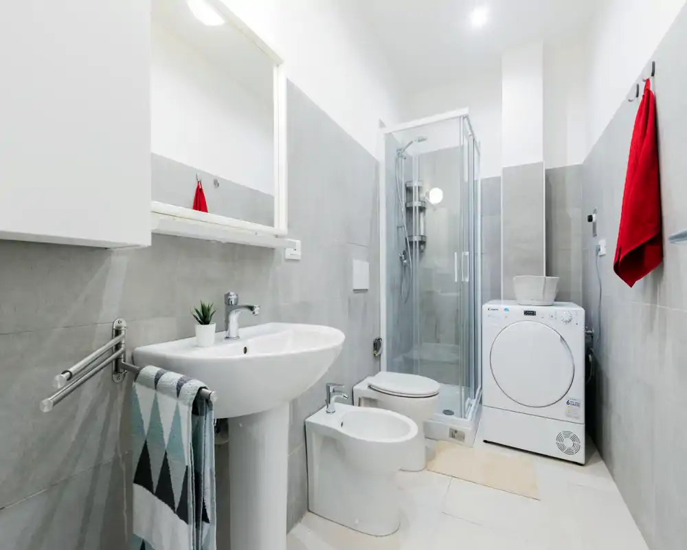 Bright, modern bathroom with a sink, bidet, toilet, glass shower and a washer/dryer; neutral grey tiles and clean white fixtures create a fresh, functional space.