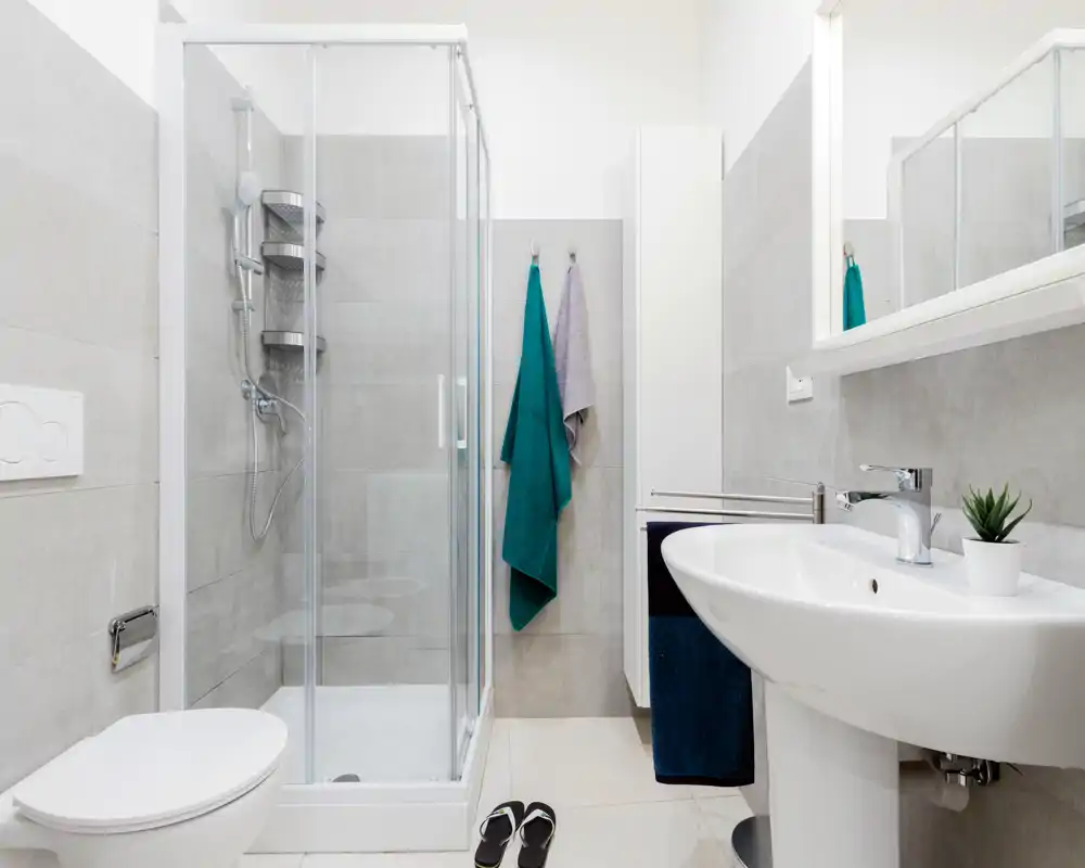 Bright, modern bathroom with a glass shower enclosure, wall-mounted toilet, and a pedestal sink. Neutral gray tiles and fresh towels create a clean, functional space.