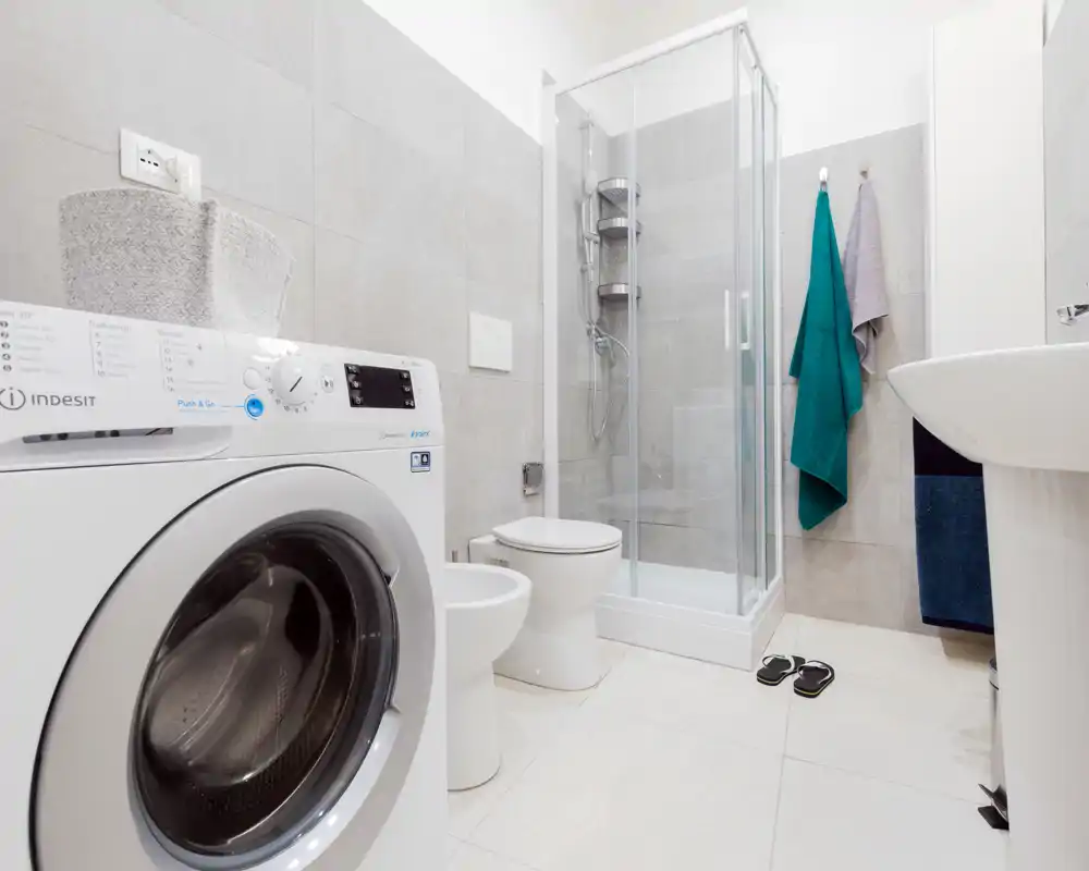Modern, bright bathroom with a glass shower enclosure, toilet and bidet, and a front-loading washing machine neatly integrated into the space.