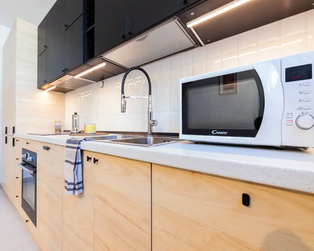 Modern kitchen with light wood cabinets, built-in oven, stainless sink and a countertop microwave; bright, clean and well-lit work surface ideal for rental photos.