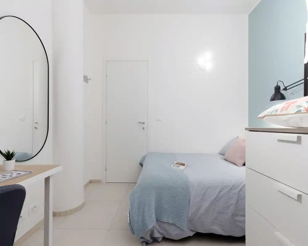 Camera da letto luminosa e minimalista con letto matrimoniale ordinato, tessuti pastello e un grande specchio ovale che amplifica la sensazione di spazio.