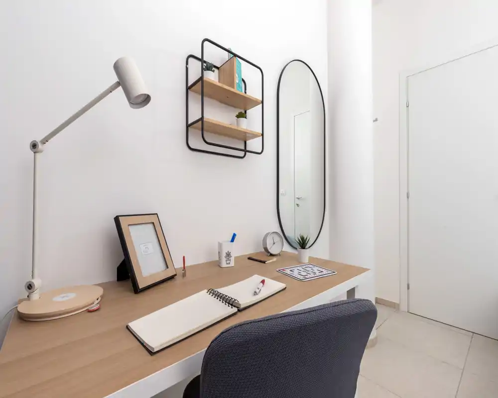 Angolo studio luminoso e minimale con scrivania in legno, lampada da lavoro, mensole a muro e specchio ovale — uno spazio compatto e ben illuminato ideale per il lavoro da remoto.