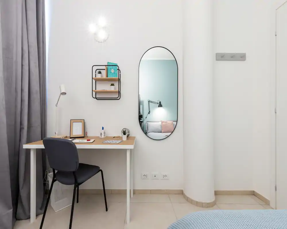 Angolo della camera da letto luminoso e moderno con una scrivania e uno specchio che riflette parte dell’area notte; stile minimalista, tessuti chiari e una lampada da comodino visibile nello specchio.