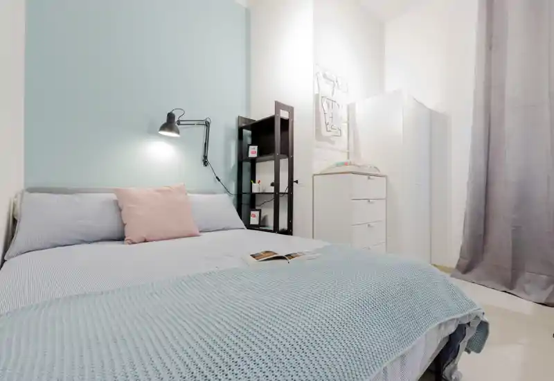 Camera da letto luminosa e moderna con un grande letto in tonalità pastello, lampada da lettura a parete e mobili contenitivi ordinati — ideale per un soggiorno riposante.