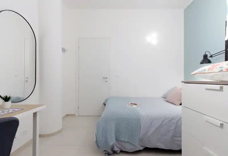 Camera da letto luminosa e minimalista con letto matrimoniale ordinato, tessuti pastello e un grande specchio ovale che amplifica la sensazione di spazio.